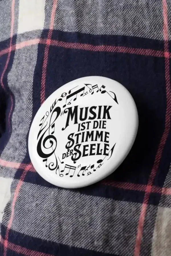 Runder weißer Musik-Button mit Noten und dem Spruch „Musik ist die Stimme der Seele“ auf kariertem Stoff.