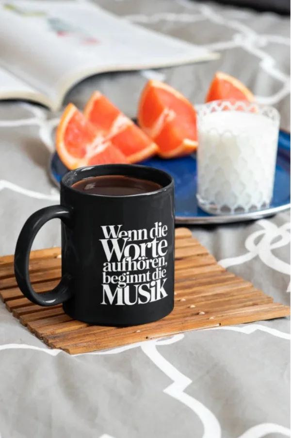 Schwarze Tasse mit dem Spruch „Wenn die Worte aufhören, beginnt die Musik“ in einer gemütlichen Frühstücksszene mit Buch und Grapefruit.