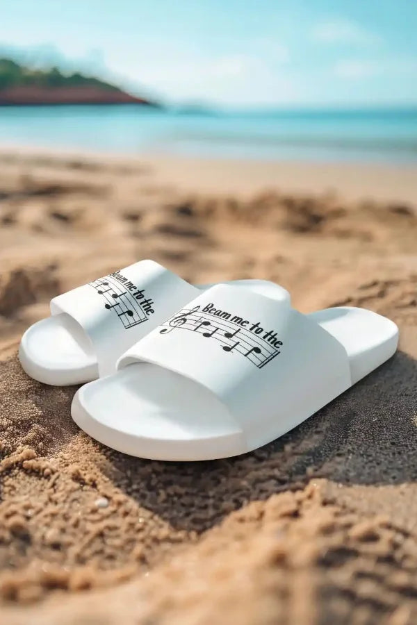 Weiße Badelatschen mit dem Notenschrift-Spruch „Beam me to the Beach“ am Strand.