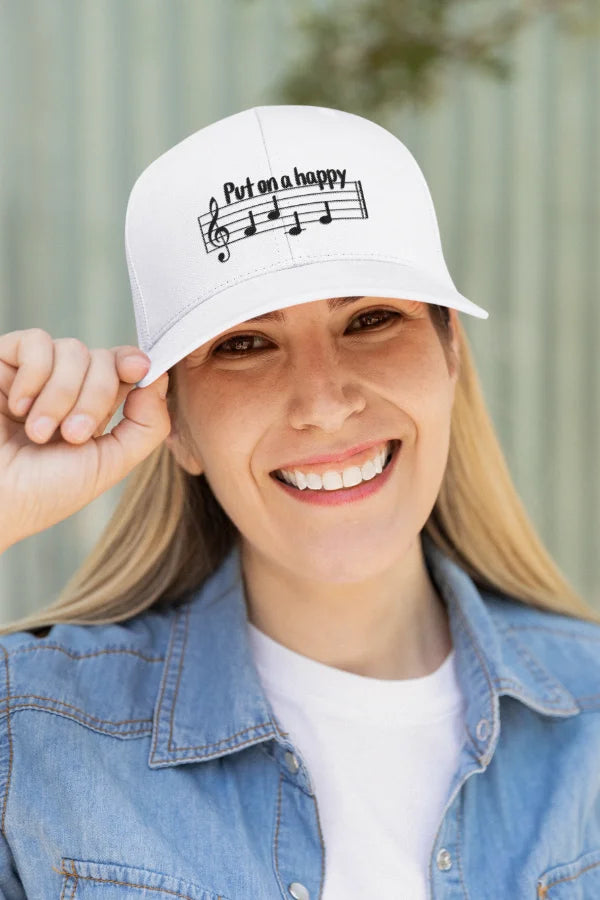 Weiße Baseball-Cap mit Notenschrift-Motiv „Put on a happy face“, getragen von einer lächelnden Frau.
