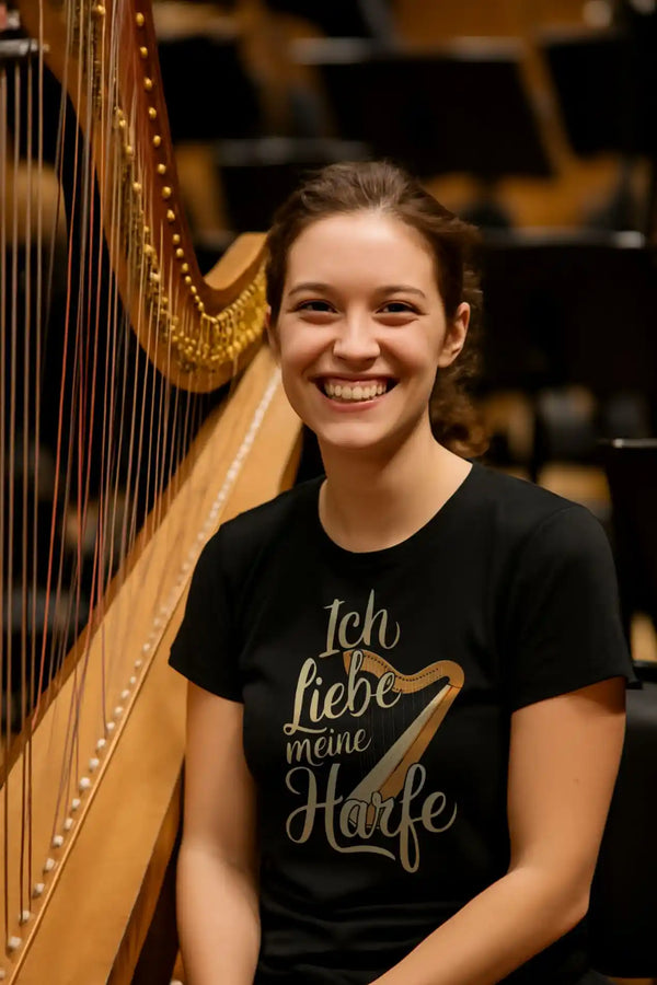 Lächelnde Harfenistin sitzt neben ihrer Harfe und trägt ein schwarzes Damen-T-Shirt mit dem Aufdruck „Ich liebe meine Harfe“