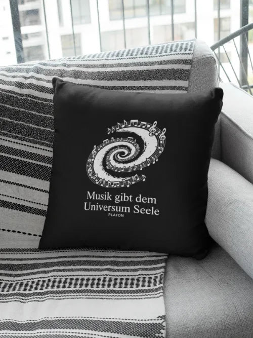 Schwarzes Kissen mit Notenspirale und dem Zitat „Musik gibt dem Universum Seele“ auf einem Sofa.