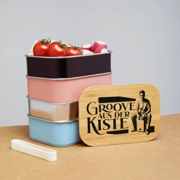 Lunchbox mit Holzdeckel und Cajón-Motiv „Groove aus der Kiste“ neben farbigen Vorratsbehältern.