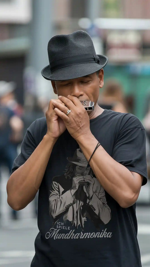 Straßenmusiker mit Hut spielt Mundharmonika und trägt ein schwarzes Herren-T-Shirt mit dem Aufdruck „Ich spiele Mundharmonika“.