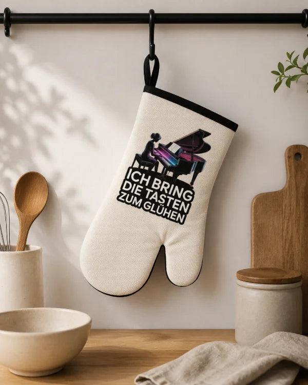 Heller Ofenhandschuh mit Klaviermotiv und Spruch „Ich bringe die Tasten zum Glühen“ in einer Küchenszene.