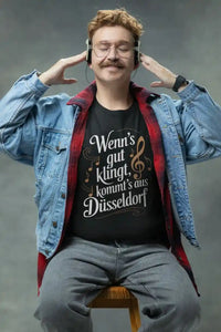 Mann mit Kopfhörern trägt ein schwarzes Herren-T-Shirt mit personalisiertem Musikdesign und dem Ortsnamen „Düsseldorf“ sowie dem Spruch „Wenn’s gut klingt, kommt’s aus Düsseldorf“.