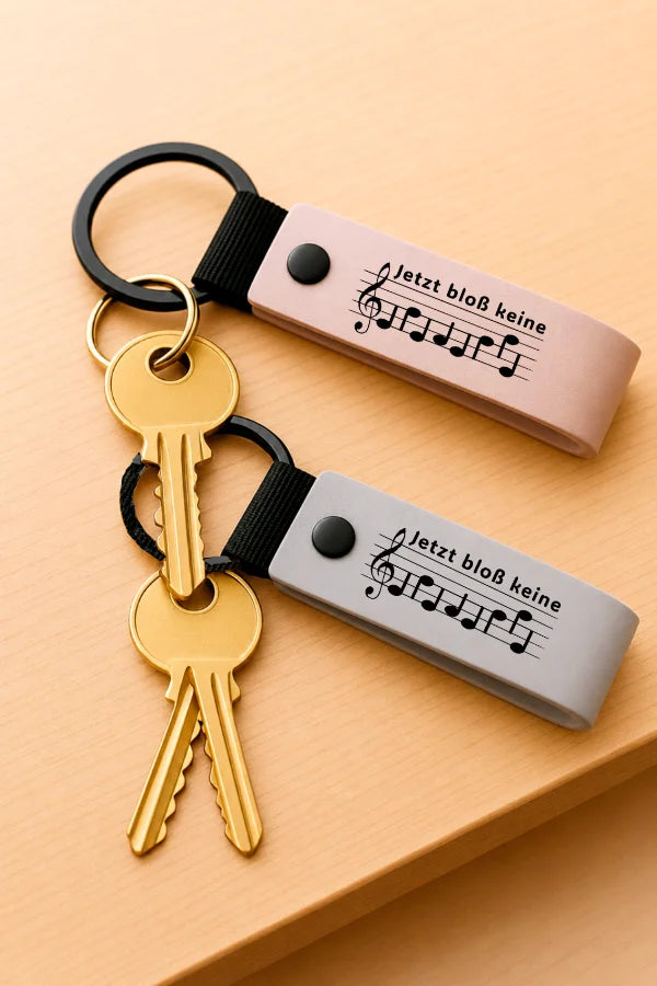 Zwei Schlüsselanhänger mit Notenschrift-Motiv „Jetzt bloß keine Ehefehde“ und Schlüsseln auf einem Holztisch.