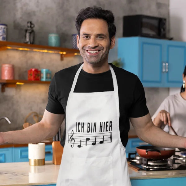 Weiße Küchenschürze mit Notenschrift-Motiv „Ich bin hier Chef“, getragen von einem lächelnden Mann in einer Küche.