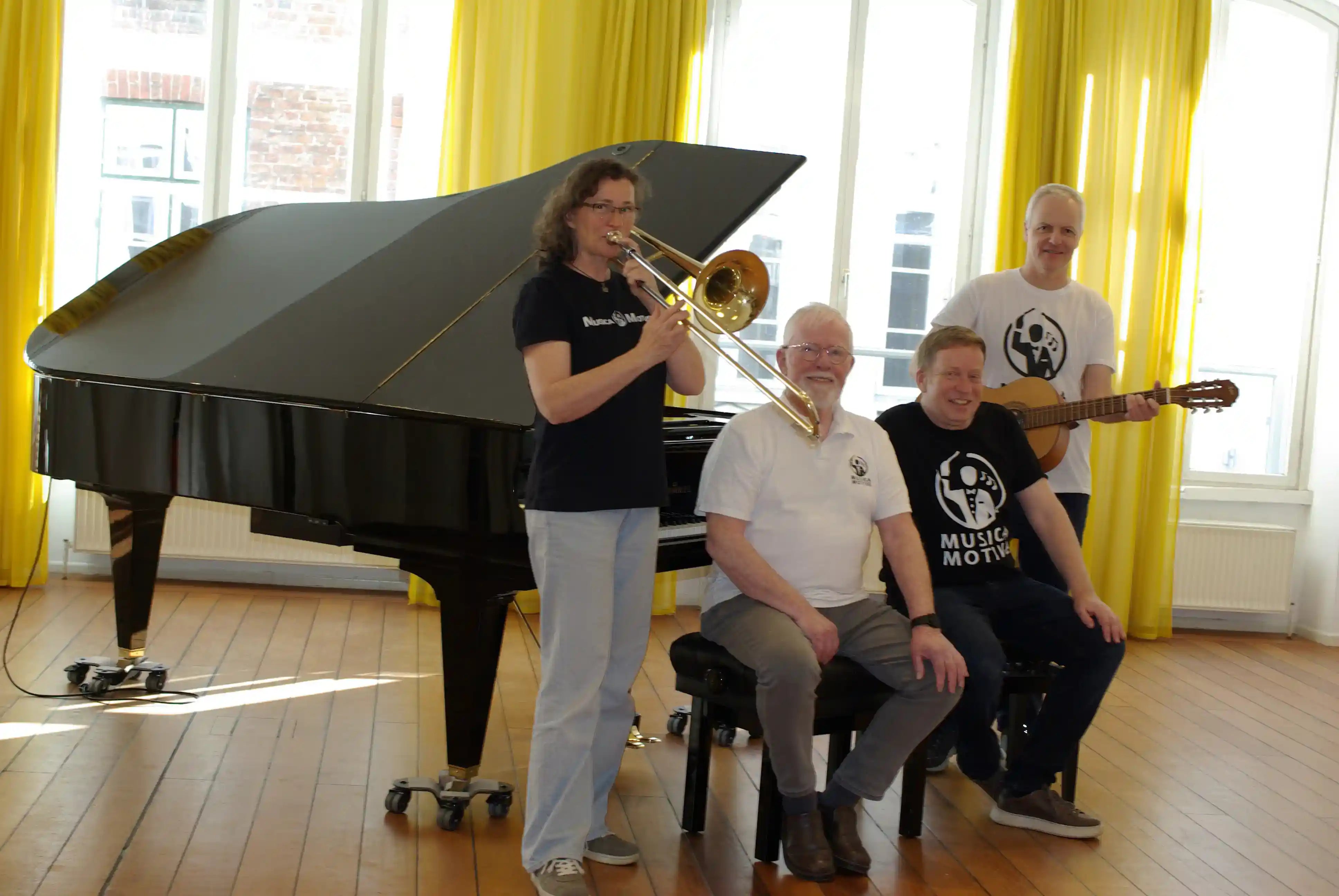 Das MusicaMotive-Team in einem hellen Musikraum mit Flügel, Posaune und Gitarre.