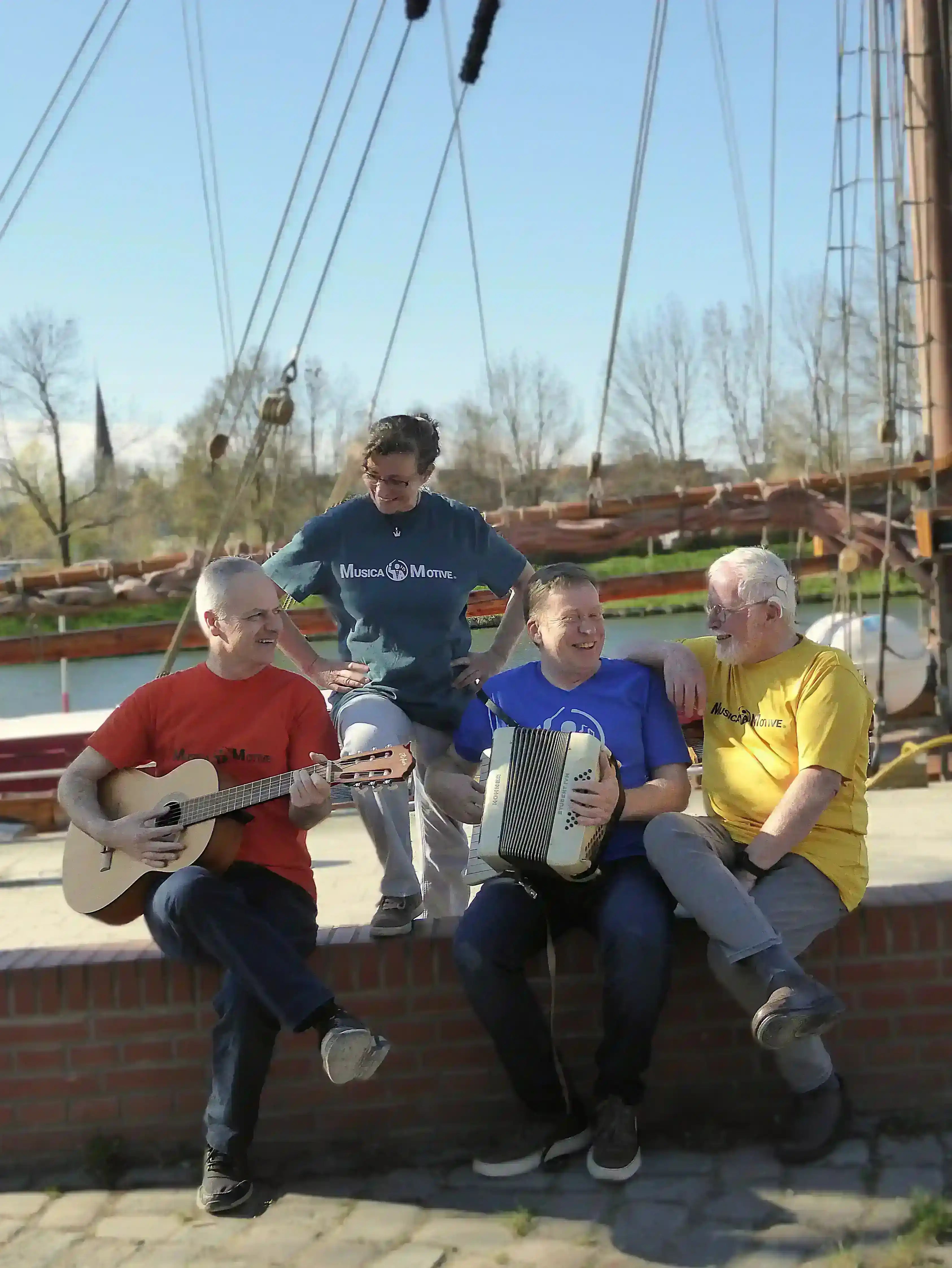 Das MusicaMotive-Team an der Trave in Lübeck mit Gitarre und Akkordeon vor einem Schiff im Hintergrund.